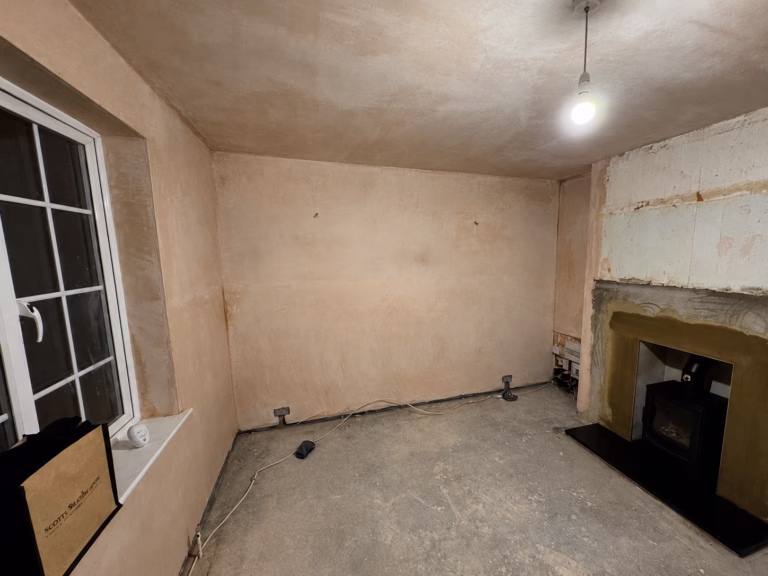 Living room before refurbishment showing window, fireplace and stripped wall surfaces awaiting renovation.